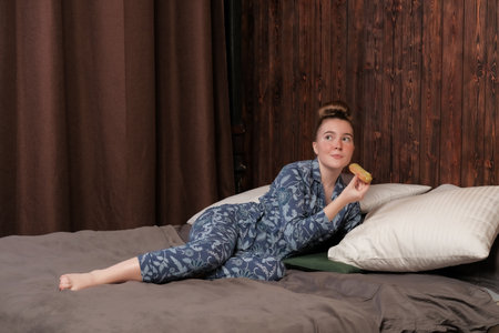 A young girl in pajamas lies on the bed and eats an eclair. Happy weekend at the hotel. Enjoyment of idleness.の写真素材