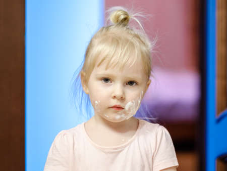 Portrait of adorable little blonde girl with cream on her face, copying the behavior of her mother, using makeup. Blurred background.の写真素材