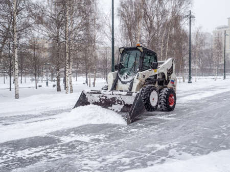 Moscow. Russia. February 12, 2021. A small loader with a bucket removes snow from a sidewalk in a...