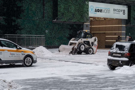 Moscow. Russia. February 12, 2021. A small loader excavator with a bucket removes snow from a road on a city street during a heavy snowfall. Winter day. Snowflakes are flying in the air.のeditorial素材