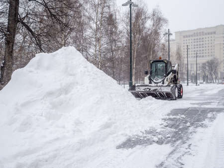 Moscow. Russia. February 12, 2021. A small loader with a bucket removes snow from a sidewalk in a city park during a heavy snowfall. Snowflakes are flying in the air. Winter day.のeditorial素材