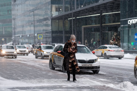 Moscow. Russia. February 12, 2021. A woman in a fur coat and a medical mask with a smartphone in her hand walks in winter along a city street against the background of many taxi cars. Snowing.のeditorial素材