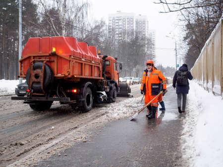Moscow. Russia. February 25, 2021. Utility workers in orange uniforms shovel muddy wet snow off a sidewalk after a heavy snowfall. Hard physical work. Cloudy winter day.のeditorial素材