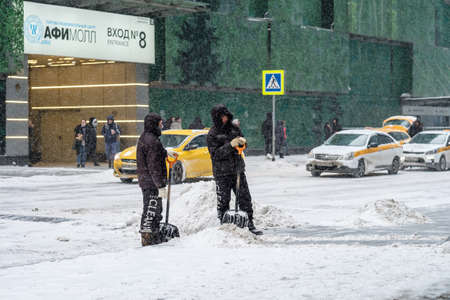 Moscow. Russia. February 12, 2021. Several utility workers with shovels remove snow from the road during heavy snowfall, with many taxi cars in the background. Snowflakes are flying in the air.のeditorial素材