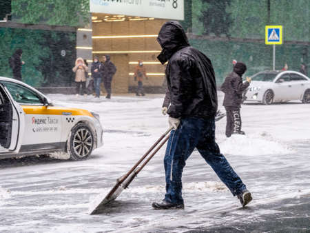 Moscow. Russia. February 12, 2021. A utility worker removes snow with a scraper on a city street during a snowfall on a winter day. Hard physical labor.のeditorial素材