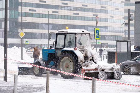 Moscow. Russia. February 12, 2021. A tractor with a bucket and a mechanical rotating brush sweeps snow on a street in a city during a snowfall. The technique is covered with a thick layer of snow.のeditorial素材