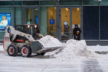 Moscow. Russia. February 12, 2021. A utility worker with a shovel and a small loader excavator remove snow from the road during a heavy snowfall. Winter day. Snowflakes are flying in the air.のeditorial素材