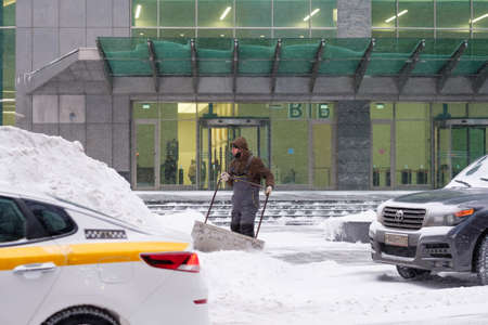 Moscow. Russia. February 12, 2021. A utility worker removes snow with a scraper on a city street during a snowfall on a winter day. Hard physical labor.のeditorial素材