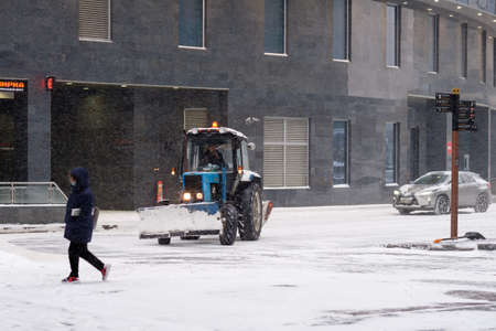 Moscow. Russia. February 12, 2021. A tractor with a bucket and a mechanical rotating brush sweeps snow on a street in a city during a snowfall. The technique is covered with a thick layer of snow.のeditorial素材