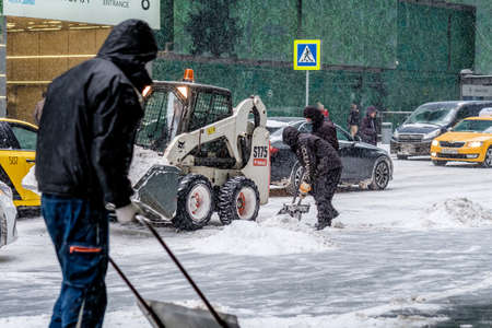 Moscow. Russia. February 12, 2021. Several utility workers with shovels and a small loader-excavator...