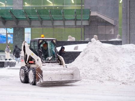 Moscow. Russia. February 12, 2021. A small loader excavator with a bucket removes snow from a road on a city street during a heavy snowfall. Winter day. Snowflakes are flying in the air.のeditorial素材