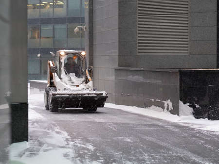 A small loader excavator with a bucket removes snow from a road on a city street during a heavy snowfall. Winter day. Snowflakes are flying in the air.のeditorial素材