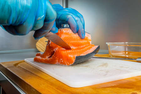 The hands of the cook in hygienic gloves with a sharp knife separate the salmon fillets from the bones. Cutting fresh fish in the kitchen on a cutting board.の写真素材