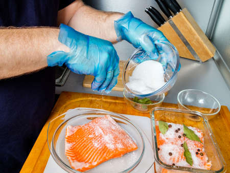The hands of the cook in hygienic gloves sprinkle salmon fillets with curing mixture. The process of cooking lightly salted fish in the kitchen.の写真素材