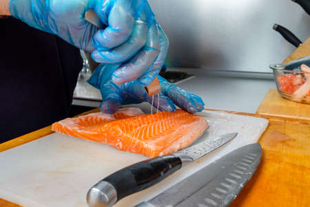A cook wearing hygienic gloves uses special fish tweezers to remove residual bones from salmon fillets. Cutting fresh fish in the kitchen on a cutting board.の写真素材