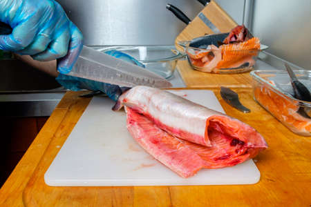 The hands of the cook in hygienic gloves cut the body of pink salmon with a knife. Cutting fresh fish in the kitchen on a cutting board.の写真素材