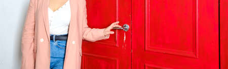 A woman in casual clothes holds on to a shiny metal handle of a vintage red wooden door. Banner. Copy space.の写真素材