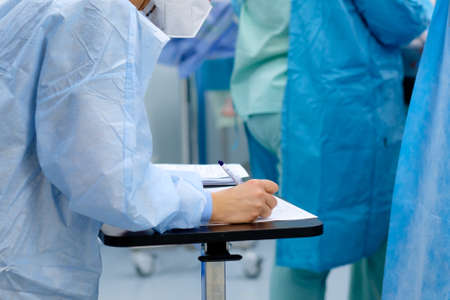 A female doctor in a blue uniform and a medical mask makes notes in documents during surgery.の写真素材