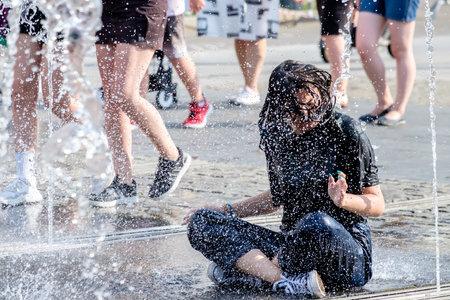 Moscow. Russia. June 26, 2021. A brunette girl in wet clothes sits on the floor of a street fountain among many drops of water on a summer day. People have fun and escape the hot weather in the city.のeditorial素材