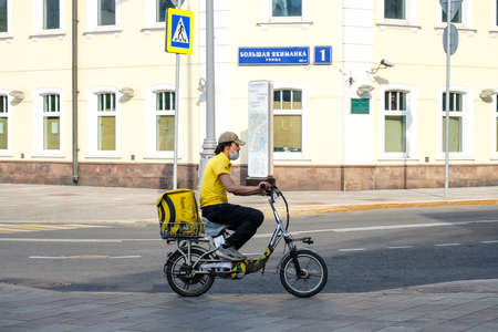 Moscow. Russia. June 26, 2021. A food delivery man wearing a protective mask rides an electric bicycle along a city street. Sunny day. Express food delivery services during the coronavirus pandemic.のeditorial素材