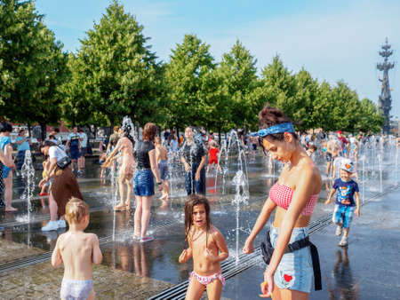 Moscow. Russia. June 26, 2021. A young pretty woman in a bikini sunbathes standing in a public park on a summer hot day. In the background, many children and adults bathe in the city fountain.のeditorial素材