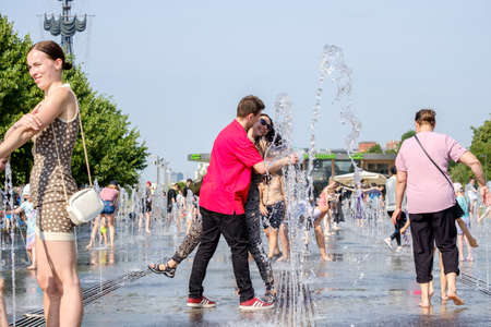 Moscow. Russia. June 26, 2021. A couple in love, a girl and a guy are smiling and hugging between splashing water in a fountain among many people. Hot summer day in the city.のeditorial素材