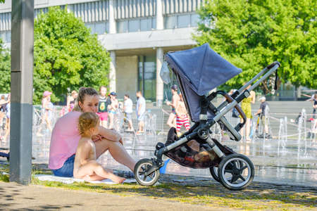 Moscow. Russia. June 26, 2021. A young mother with a small child sits on the ground near the city fountain. Walk outdoors on a sunny summer day. There is a baby stroller nearby.のeditorial素材