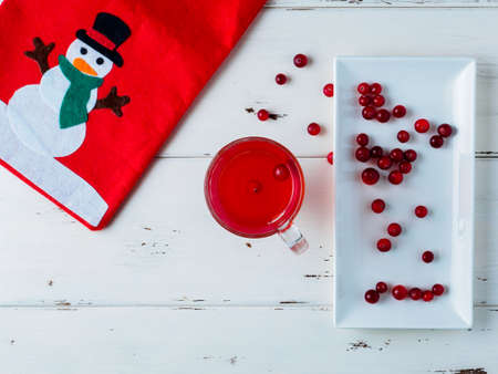 Selective focus on cranberries in a fresh drink in a glass cup. Berries on a white plate. Red bag with a picture of a snowman. New Years decor on a white wooden background. Copy space. Flat lay.の写真素材