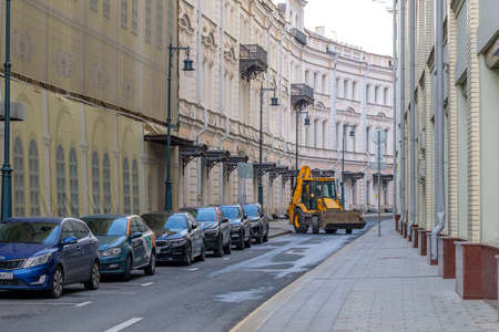 Moscow. Russia. August 1, 2021. A yellow tractor with a bucket drives down a narrow street in the city center along parked cars. Work of municipal equipment.のeditorial素材