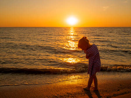 Silhouette of a little child girl in a striped dress on the seashore against the backdrop of the setting sun.の写真素材