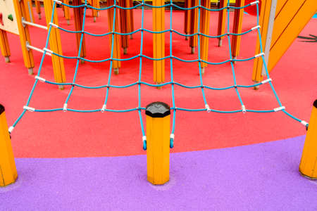 A climbing net, woven of ropes, on a playground against a brightly colored rubberized floor. Red and...