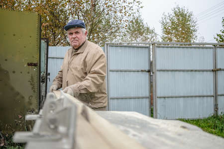 An elderly grey-haired carpenter cuts a board on a machine with a circular saw. Selective focus. A...