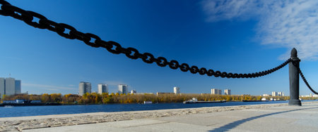 A fragment of a cast-iron chain fence on the river embankment. In the background, water, blue skies and high-rise buildings on the opposite bank of the river. Sunny day. Panorama.の写真素材