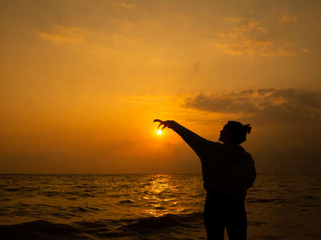 Silhouette of a young woman on the seashore at sunset. The girl touches the sun with her fingers. Orange disk of the sun in the hands of a girl.の写真素材