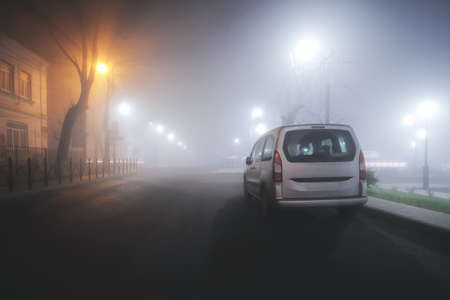 Silver minivan in dense fog on a night street in the city. Soft focus. The light of street lamps along the road in the fog. Car on a foggy autumn night in the city.の写真素材