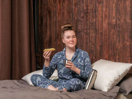 A young girl in pajamas sits on the bed and eats an eclair. Hot tea from a flask. Happy weekend at the hotel.の写真素材