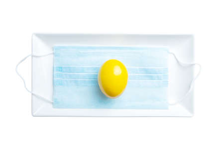 Isolated image of a yellow egg lying on a medical mask inside a white rectangular plate on a white background. The concept of protecting life with self-isolation during the coronavirus epidemic.の写真素材