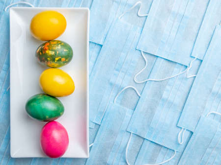 Colored Easter eggs on a white rectangular ceramic plate on a background of blue medical masks. COVID-19 Flat lay. Easter holiday concept during quarantine coronavirus epidemic. Copy spaceの写真素材