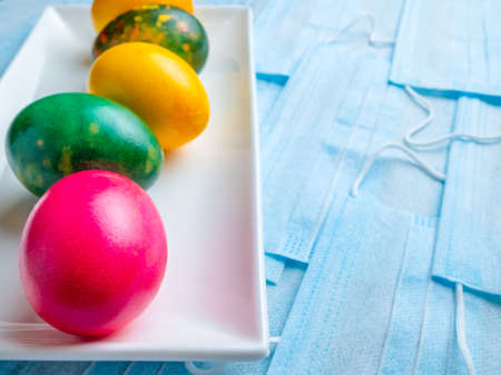 Colored Easter eggs on a white rectangular ceramic plate on a background of blue medical masks. COVID-19 Flat lay. Easter holiday concept during quarantine coronavirus epidemic. Copy spaceの写真素材