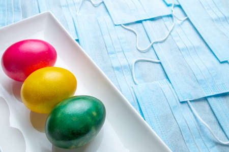 Colored Easter eggs on a white rectangular ceramic plate on a background of blue medical masks. COVID-19 Flat lay. Easter holiday concept during quarantine coronavirus epidemic. Copy spaceの写真素材