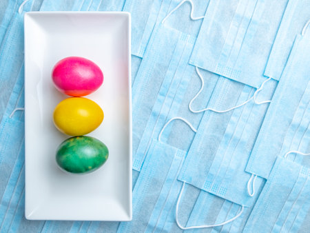Colored Easter eggs on a white rectangular ceramic plate on a background of blue medical masks. COVID-19 Flat lay. Easter holiday concept during quarantine coronavirus epidemic. Copy spaceの写真素材