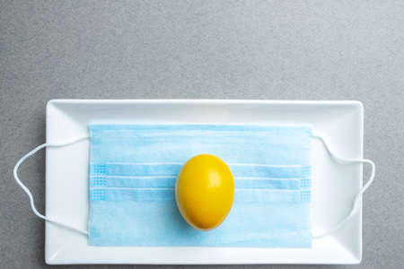 Yellow easter egg lies on a blue medical mask inside a white rectangular ceramic plate on a gray background. Flat lay. Easter holiday concept during quarantine coronavirus epidemic. Copy space.の写真素材