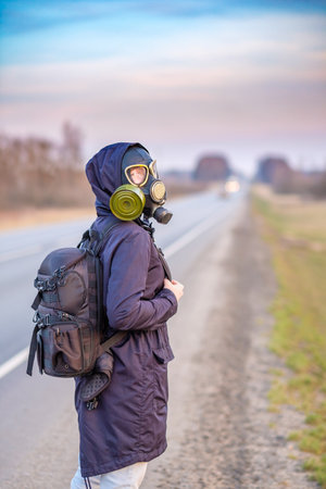 A girl in a gas mask hitchhikes and walks along the side of the road past passing cars. Escape from the area infected with the coronavirus.の写真素材