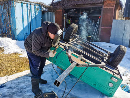 An elderly male welder repairs a car trailer with electric arc welding on a spring sunny day.の写真素材