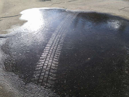 Track from a studded tire on the ice of a frozen puddle on cracked asphalt in the bright sunshine on...