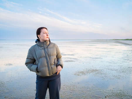 Young woman by the sea on a cold day at low tide. Portrait of a young girl on the beach during sunset on a cold evening.の写真素材