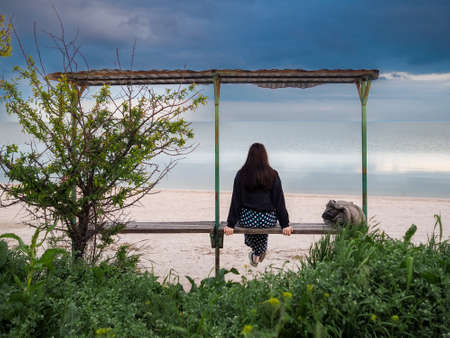 Rear view of a woman sitting on a bench in front of the sea. Lonely young woman sits on a bench by the sea on a cloudy day.の写真素材
