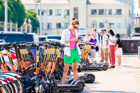 Moscow. Russia. June 9, 2022. A young woman rents an electric scooter using an app in her smartphone on a summer sunny day. Selective focus. Public eco-friendly electric transport in the city.のeditorial素材