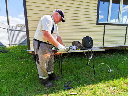 An elderly man cuts metal pipes with a disc grinder. The master works as a grinder in the yard of a country house on a sunny summer day.の写真素材
