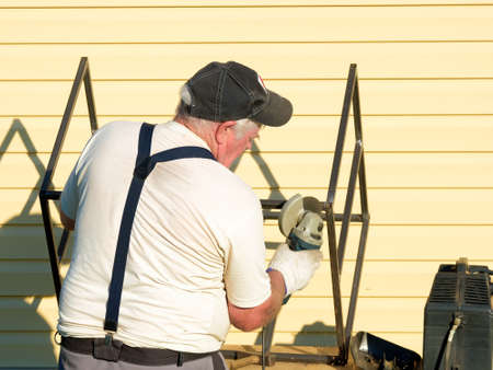 An senior gray-haired man processes a metal structure with a disc grinder. The master processes metal in the yard of a country house on a sunny summer day.の写真素材